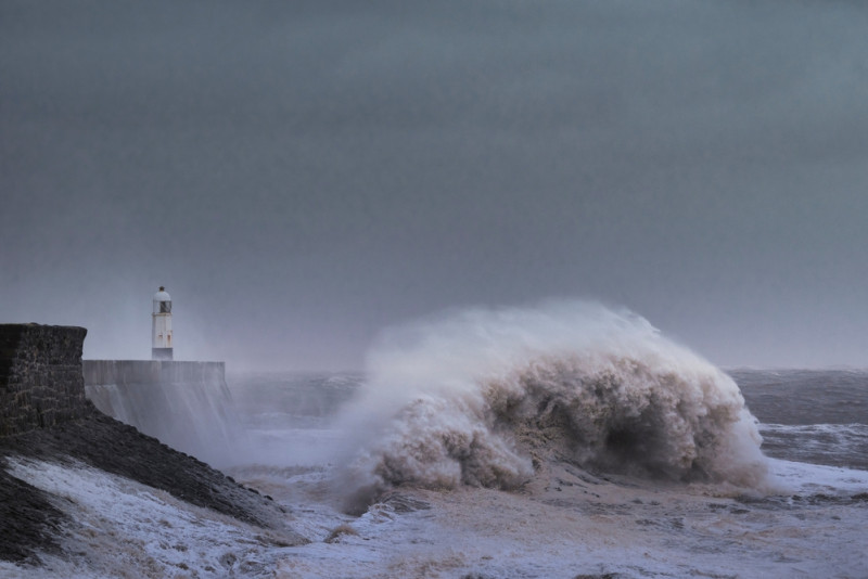Une vague s'écrase sur une digue devant un phare2025
