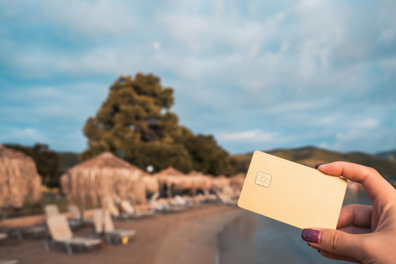Une personne tient une carte bancaire devant un paysage de vacances