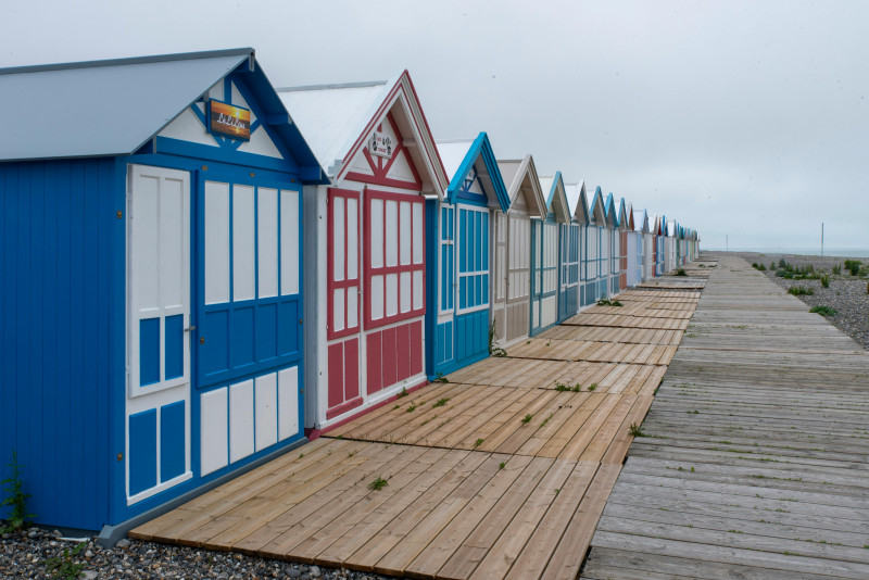 Des cabines de plage colorées bordent la promenade de Cayeux-sur-Mer, en France, sous un ciel nuageux.