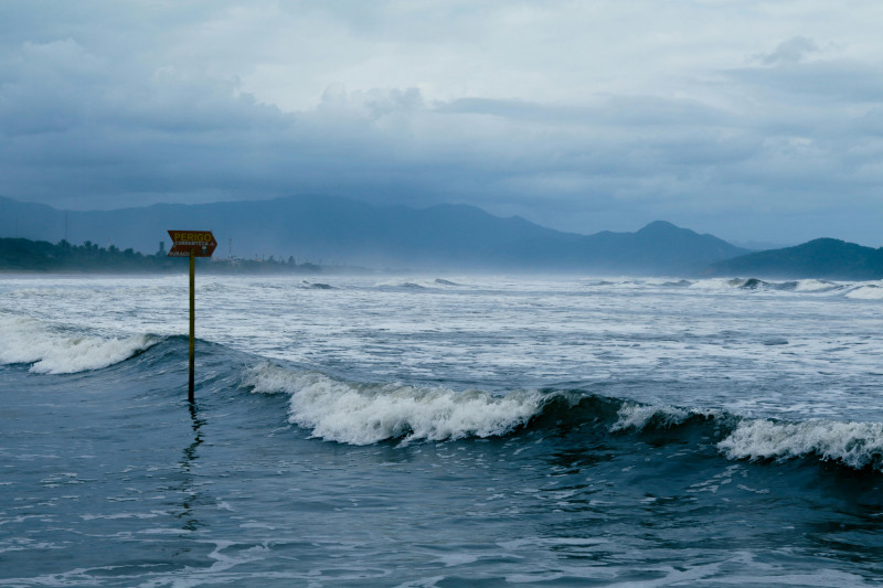 Scène côtière nuageuse avec un panneau d'avertissement et des vagues s'écrasant contre le rivage, transmettant la prudence en mer.