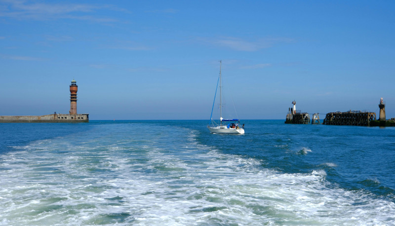 Un paysage marin serein avec voilier et phare sous un ciel bleu à Dunkerque, France.