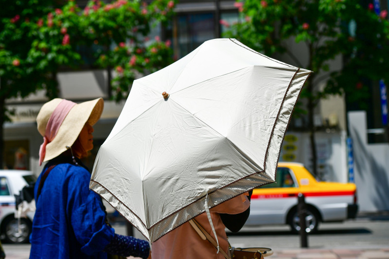 A close-up street style photograph of women shielding themselves from the bright summer sun with a white parasol umbrella and a straw hat on a busy city street in Japan. 📸 Photo by me IG: @margoevardson