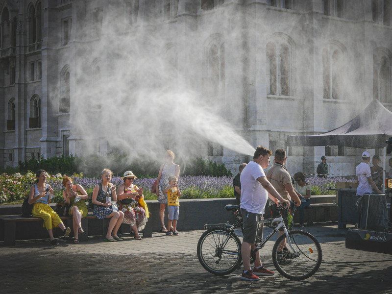 Street scene on a hot summer day in Budapest with a cyclist passing through a cooling mist, people sitting on benches and enjoying the shade. A humorous and refreshing moment of everyday life.