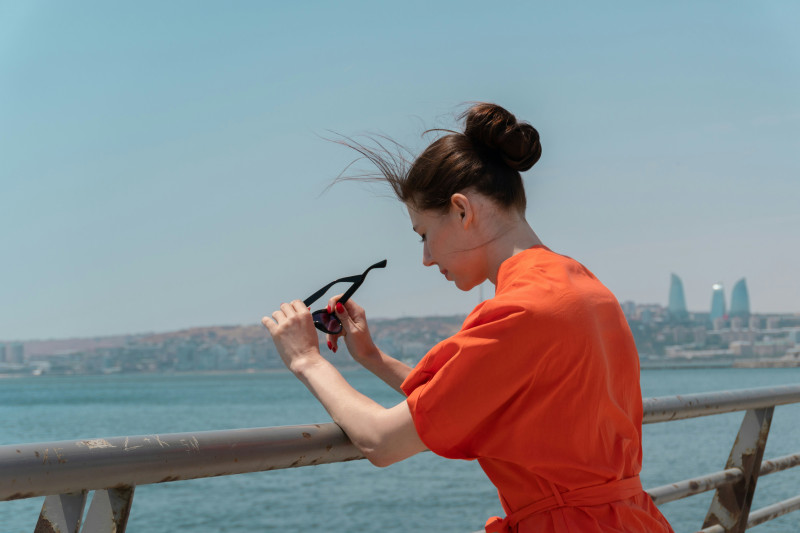 Woman in orange dress holding sunglasses by the water.
