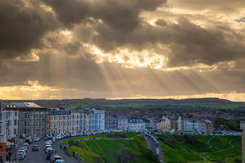 Des rayons de soleil percent à travers des nuages dramatiques sur un paysage urbain côtier avec des bâtiments historiques.