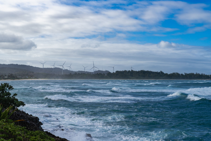 Vue oceanique panoramique avec de puissantes vagues et des éoliennes au loin sous un ciel nuageux.