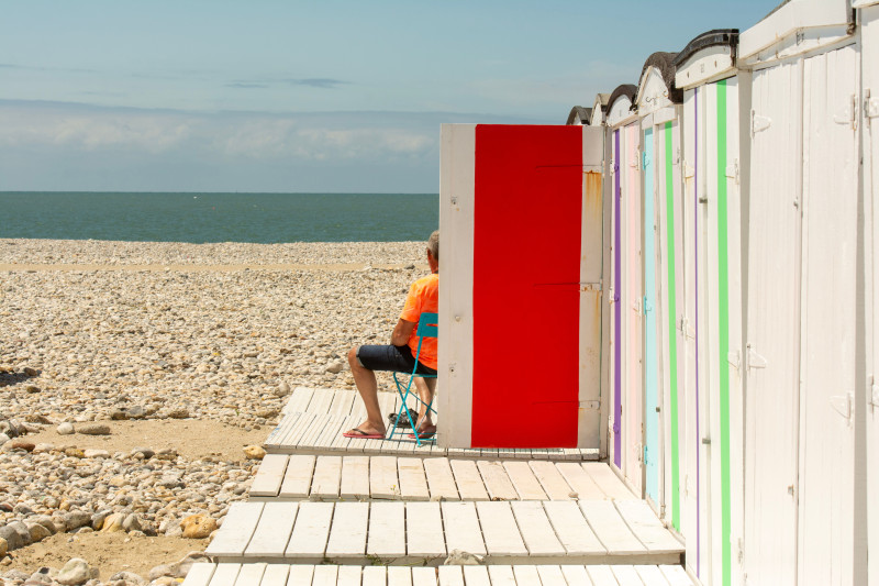 Personne assise près de cabanes de plage colorées sur une plage de galets, profitant d'une vue sereine sur l'océan.