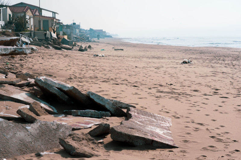 Une scène côtière sereine avec du béton cassé le long d'une plage de sable et des maisons à l'arrière-plan.