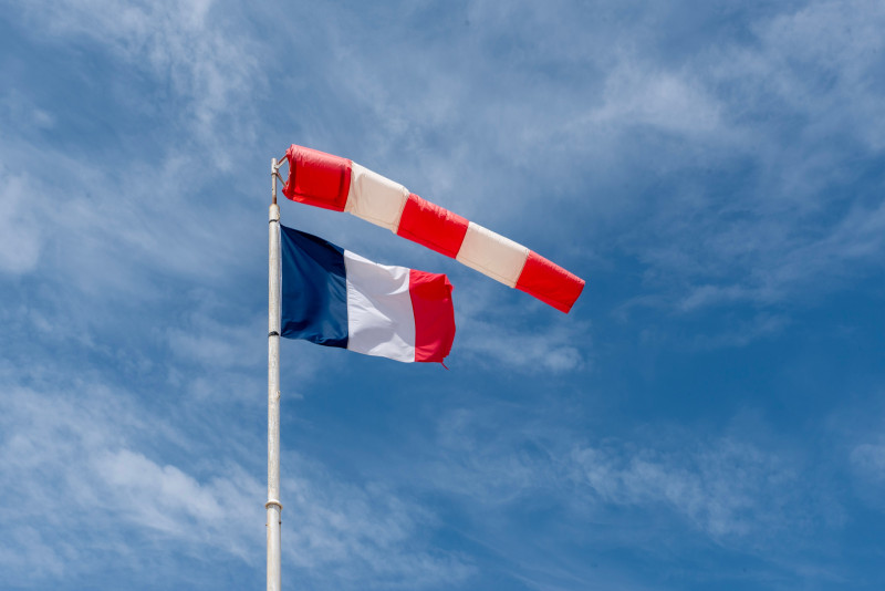 Drapeau français et manche à air rouge et blanche flottant sous un ciel bleu parsemé de nuages épars.