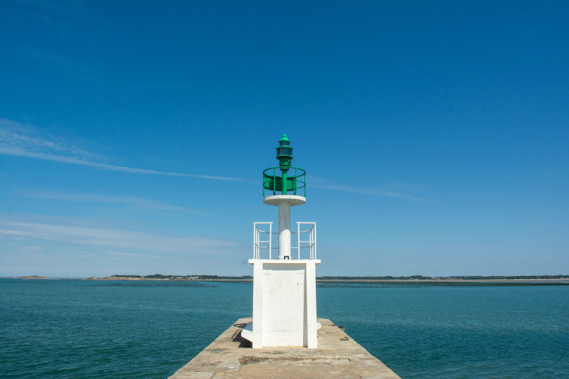Vue sereine d'un phare sur une jetée s'étendant dans la mer à Mesquer, France, sous un ciel bleu dégagé.