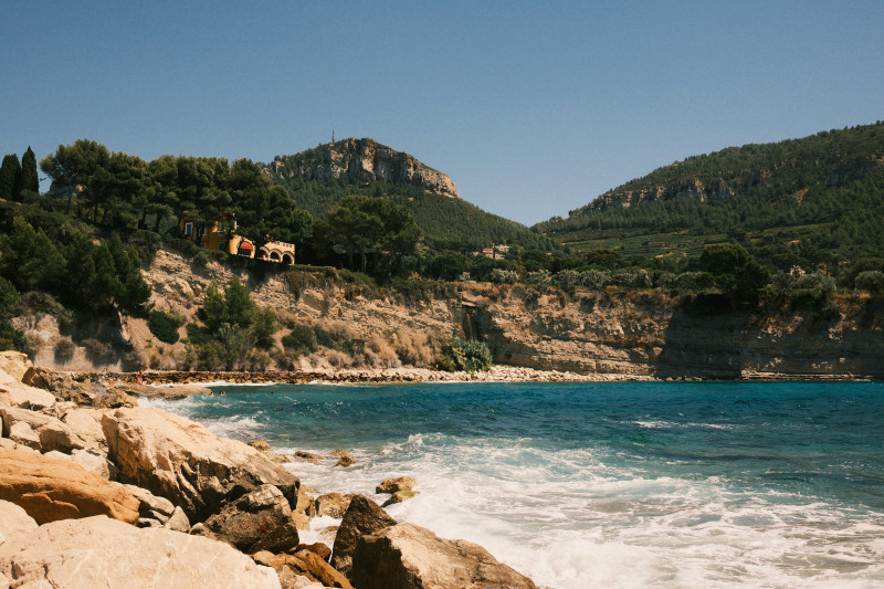 A scenic view of the rocky coastline near Cassis, in the Provence-Alpes-Côte d’Azur region of France. The turquoise Mediterranean Sea crashes against the cliffs under a clear blue summer sky, with pine trees and hillside villas overlooking the water. Perf