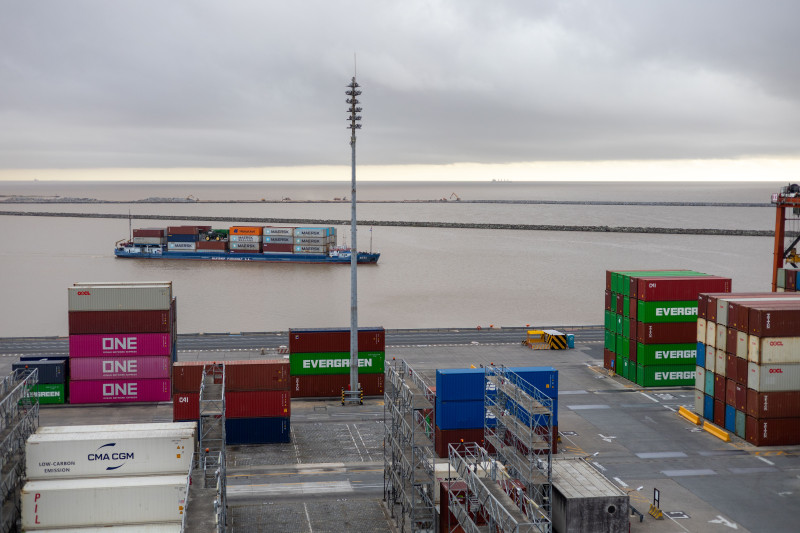 Un porte-conteneurs dans un port, entouré de conteneurs. Journée nuageuse.