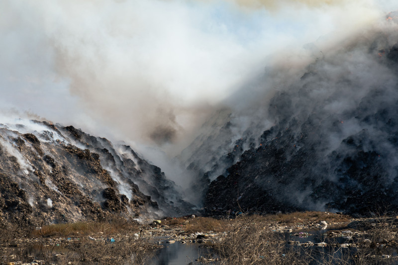 Une dense fumée s'échappe d'une décharge en feu entourée de tas de déchets.