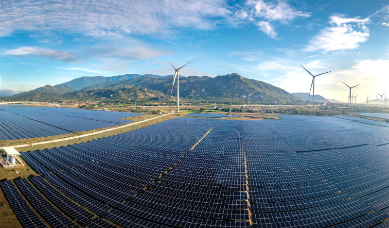 Vue aérienne d'une vaste ferme solaire avec des éoliennes sur fond de montagnes par une journée ensoleillée.