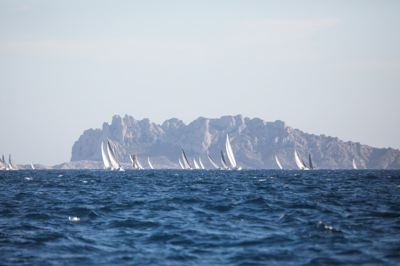Au loin, des voiliers fendent les eaux d'un bleu éclatant au large de Marseille, avec des falaises rocheuses en arrière-plan.