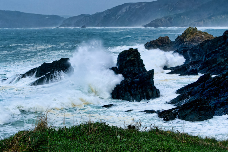 Waves crashing against rugged rocks along a coastal landscape during a stormy afternoon in a remote location - Powerful waves surge against dark, jagged rocks while foam froths on the water's surface. The dramatic coastline exhibits wild, natural beauty, 