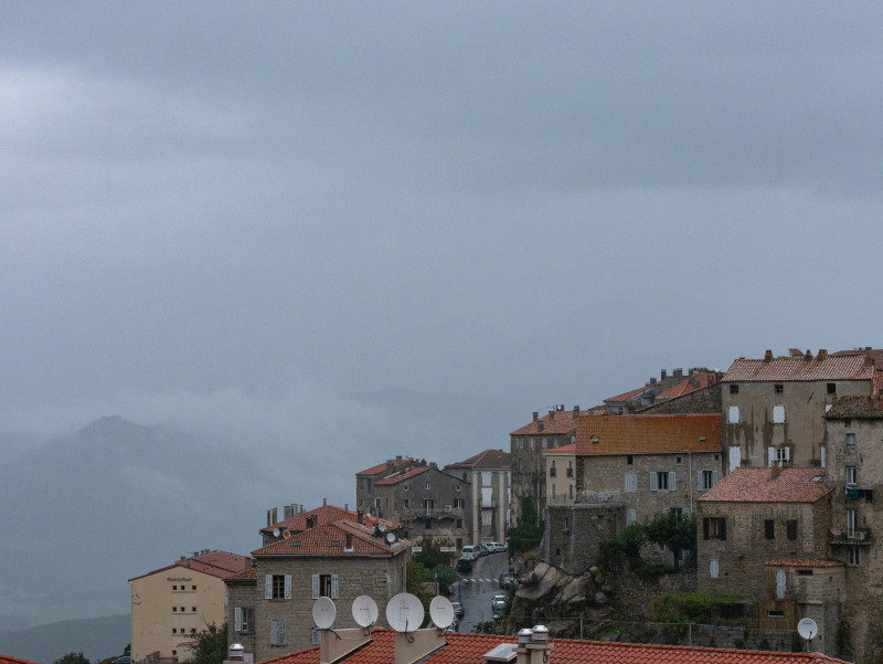 A picturesque view of a Corsican town nestled between misty mountains, where charming buildings with warm-toned facades create a striking contrast against the dramatic, fog-covered peaks. The blend of urban and natural elements, with winding streets and s