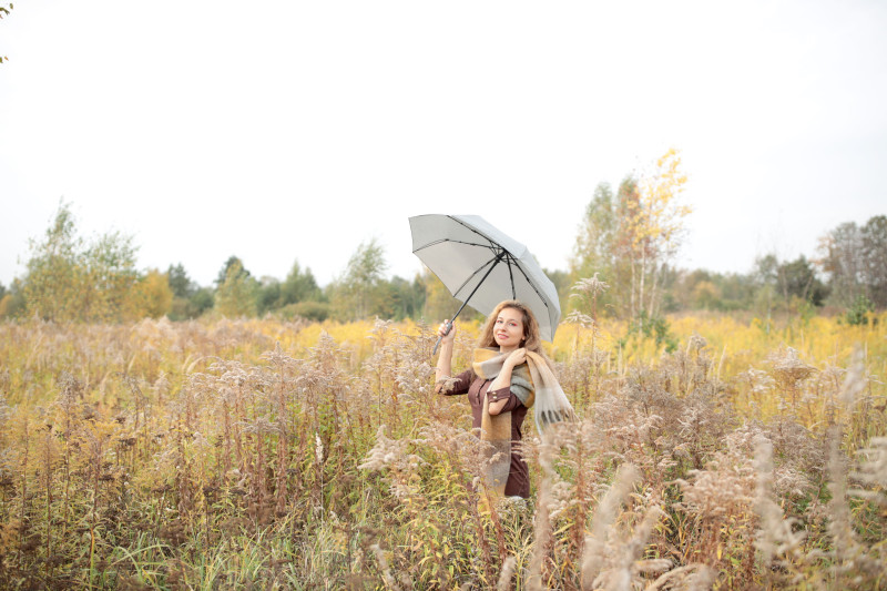 Une femme se tient dans un champ d'automne tenant un parapluie, entourée de feuillage d'automne.