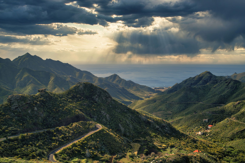 Vue à couper le souffle sur une chaîne de montagnes avec une route sinueuse sous un ciel dramatique et des rayons de soleil.